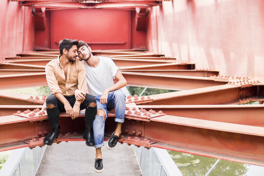 Young gay couple in love sitting on steel girder of a footbridge