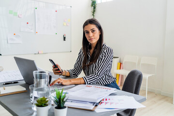Confident female design professional sitting with wireless technologies at desk in office