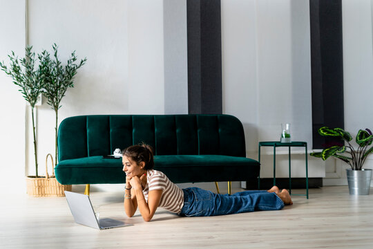 Smiling businesswoman looking at laptop while lying on floor by sofa in creative workplace