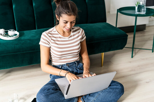 Smiling businesswoman using laptop while sitting cross-legged on floor against sofa at office