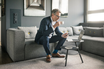 Mature businessman with cup of coffee and laptop using cell phone on couch