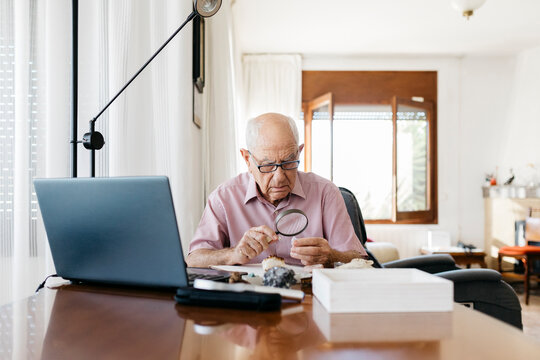 Retired elderly man analyzing minerals and fossils with magnifying glass while sitting at table