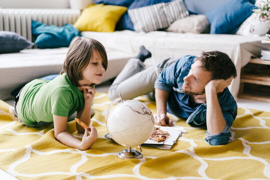 Father and son eating pizza next to globe on the floor at home