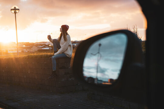 Iceland, young woman with coffee to go at sunset, wing mirror