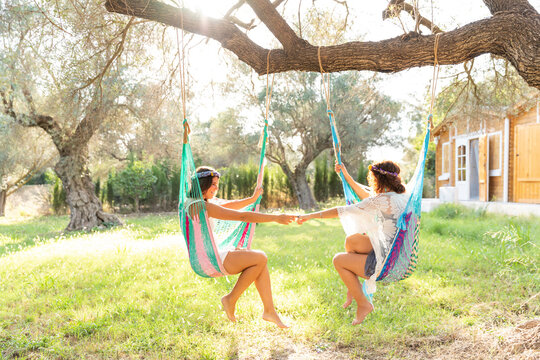 Young women holding hands while sitting in hammocks at yard