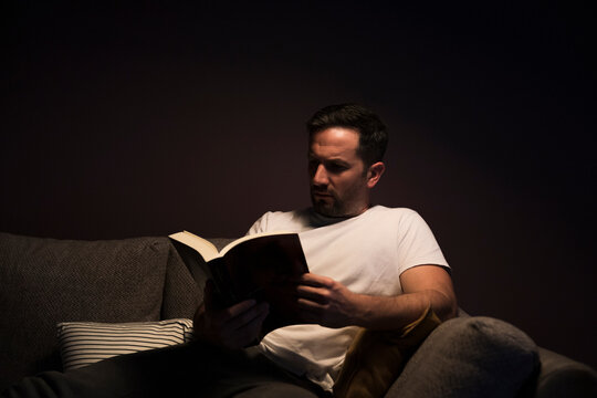 Mature man reading book while sitting on sofa against wall at home