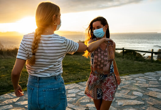 Female friends in protective face masks elbow bumping on footpath during COVID-19 outbreak