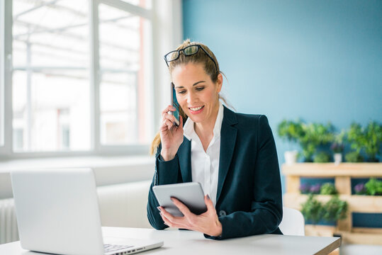 Businesswoman sitting at desk, talking on the phone, looking at digital tablet