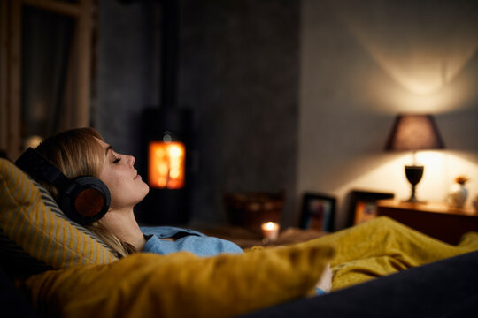 Smiling woman listening music with headphones on couch at home in the evening