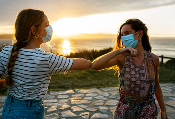 Women in protective face masks elbow bumping against sky during COVID-19 outbreak - obrazy, fototapety, plakaty