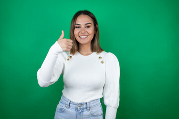 Young beautiful woman wearing white sweater over isolated green background smiling and doing the ok signal with her thumb