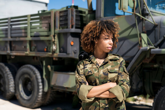Young female army soldier with arms crossed looking away while standing against truck at military base