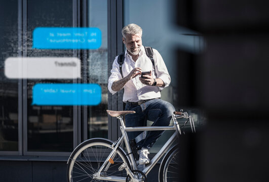 Smiling mature businessman with bicycle using cell phone for messaging in the city
