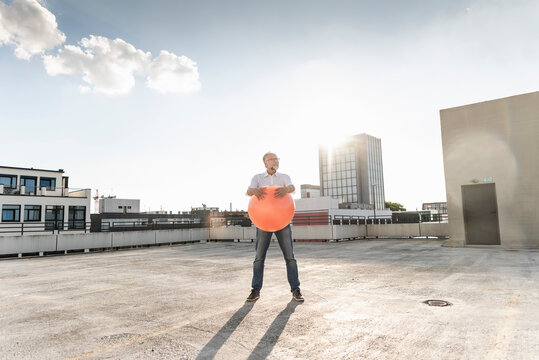 Mature man playing with orange fitness ball on rooftop of a high-rise building