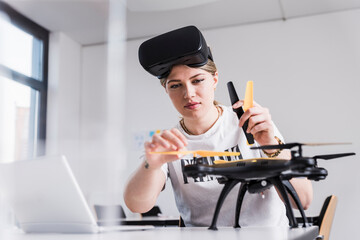 Young woman with laptop and VR glasses at desk examining drone