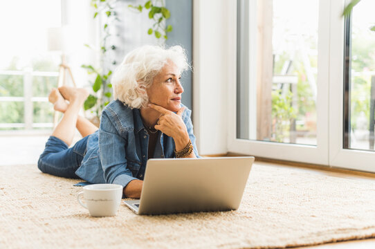 Mature woman lying on front while looking away working on laptop at home