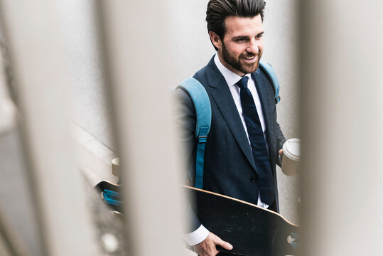 Smiling businessman with takeaway coffee and skateboard walking outdoors