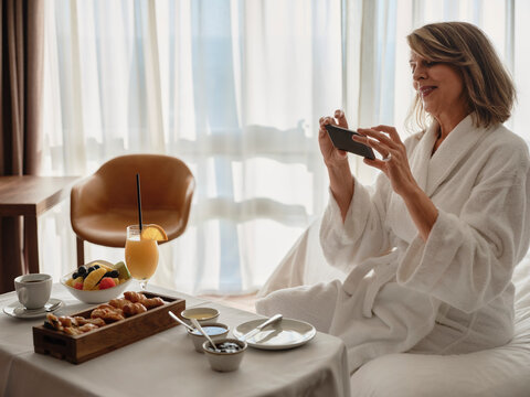 Smiling blond elderly woman photographing breakfast through smart phone while sitting on bed in hotel room