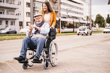 Smiling young woman pushing happy senior man with smartphone in wheelchair