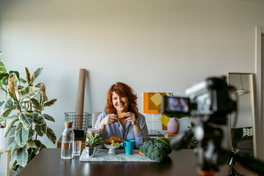 Smiling woman showing fresh fruits while making vlog on camera at home