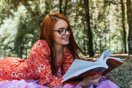 Smiling young beautiful redhead woman reading book while lying on picnic blanket with diary at park