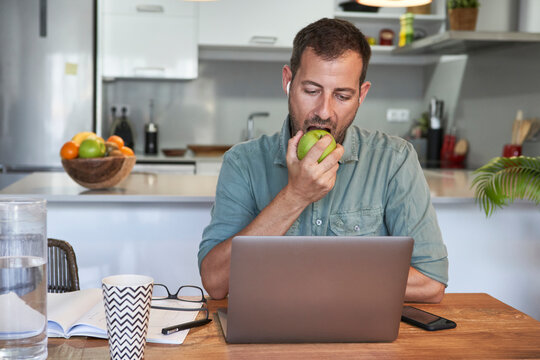 Handsome male freelancer eating fresh apple while working at home with laptop