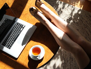 Woman with her legs up on desk with a herbal tea and laptop with sun streaming in window