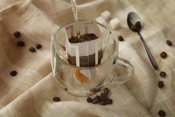 Pouring hot water into glass cup with drip coffee bag on beige fabric, closeup