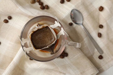 Glass cup with drip coffee bag and beans on beige fabric, flat lay