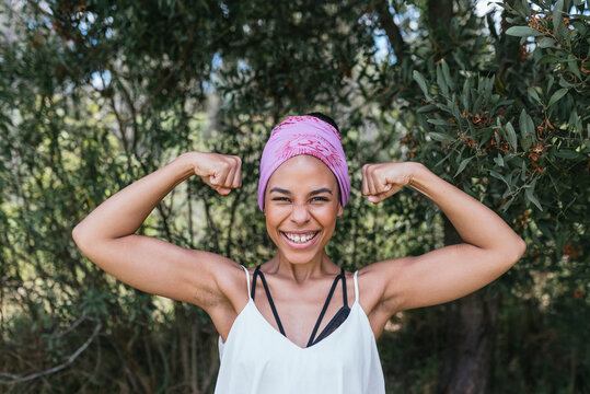 Happy woman with purple bandana flexing muscles while standing against plants at park