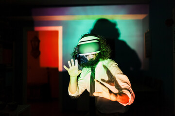 Smiling man wearing virtual reality glasses standing in multi colored light reflection at home during pandemic