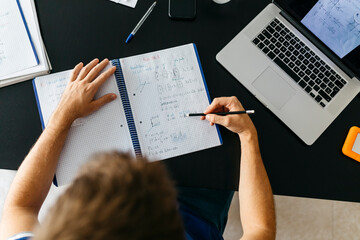 Male student solving mathematical equations in workbook while sitting at table