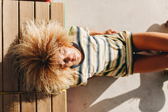 Woman with blond curly hairstyle with eyes closed reclining on wooden seat during sunny day