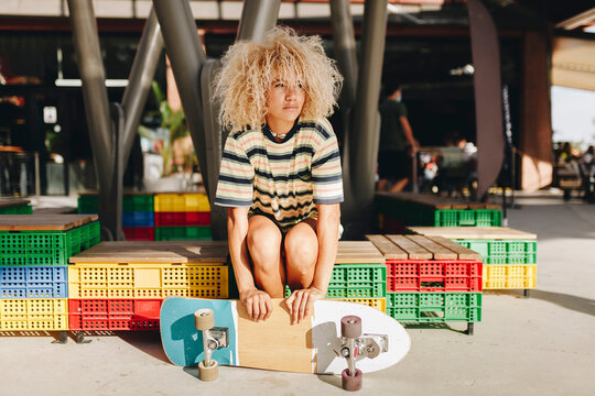 Afro blond woman holding skateboard and looking away while sitting on crate seat during sunny day