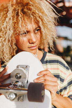 Hispanic woman with blond curly hair looking away while holding skateboard on sunny day