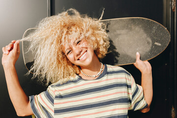 Cheerful woman holding skateboard while pulling curly blond hair against wall