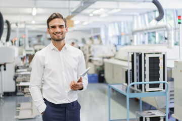 Smiling confident young businessman holding digital tablet while standing with hands in pockets at illuminated factory