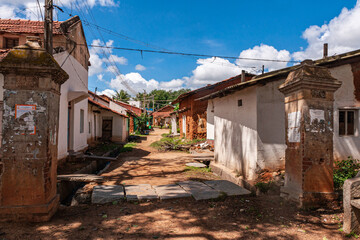 Fototapeta premium Kadenahalli, Karnataka, India - November 3, 2013: Looking into the main street of the rural village under blue cloudscape. Dirt road with open sewer and short houses in white and red. Green foliage in