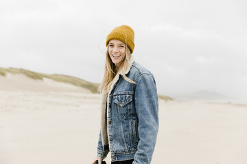 young woman wearing yellow beanie and denim jacket on empty beach smiling toward camera