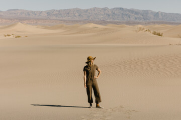 young woman in fashionable green jumpsuit and hat standing on sand dunes with mountains in background