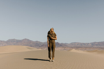 young woman in fashionable green jumpsuit and hat standing on sand dunes with mountains in background