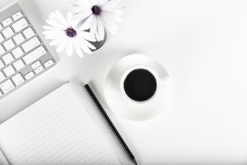 White desk with white flowers in a vase, note pad, pencil, keyboard, and coffee in a white coffee mug. Directly above view.