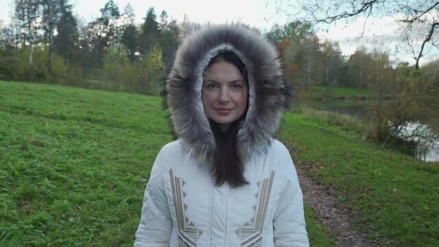 Beautiful woman with brown hair walking in park on cold autumn day with trees covered in yellow leaves in warm fur collar coat
