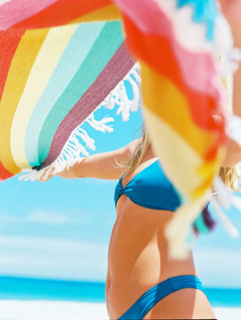 Blonde girl in blue bikini at sunshiney beach in summer with sunglasses and rainbow towel