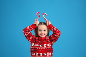 Cute little girl in knitted Christmas sweater holding candy canes on blue background