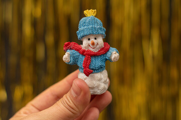 Woman hand holding a happy snowman figurine with a warm hat, scarf and sweater on golden...