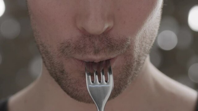 Close-up side view of young unidentified european man eating a juicy piece of meat from a fork on a shiny background. Healthy wholesome food concept. Slow motion
