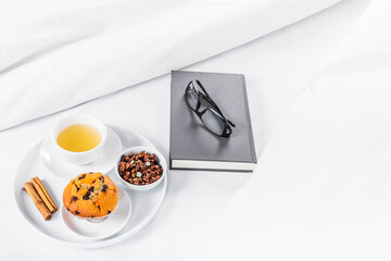Breakfast tray on the bed, with tea, muffin, cinnamon and granola, white sheets and grey book and eyeglasses. White Background, copy space.