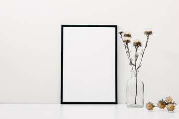 Black photo frame and glass vase with dried flowers on white background. front view