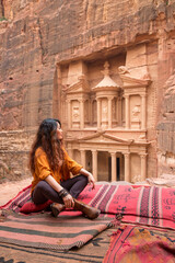A beautiful girl in an orange shirt sits cross-legged on the edge of a rock and looks at El-khazneh